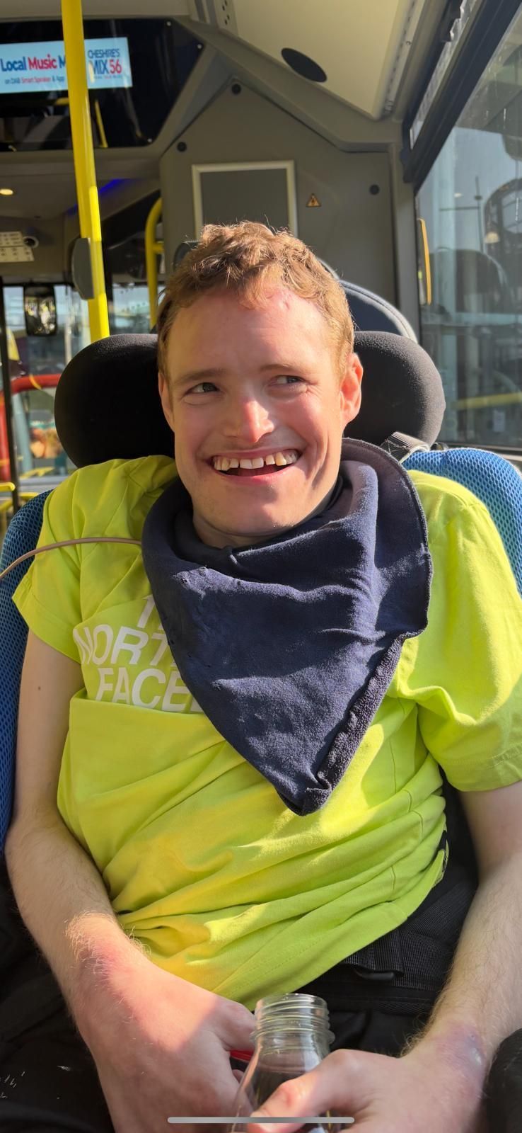 Man with curly hair on a bus, wearing a yellow shirt and blue scarf, smiling and holding a bottle.