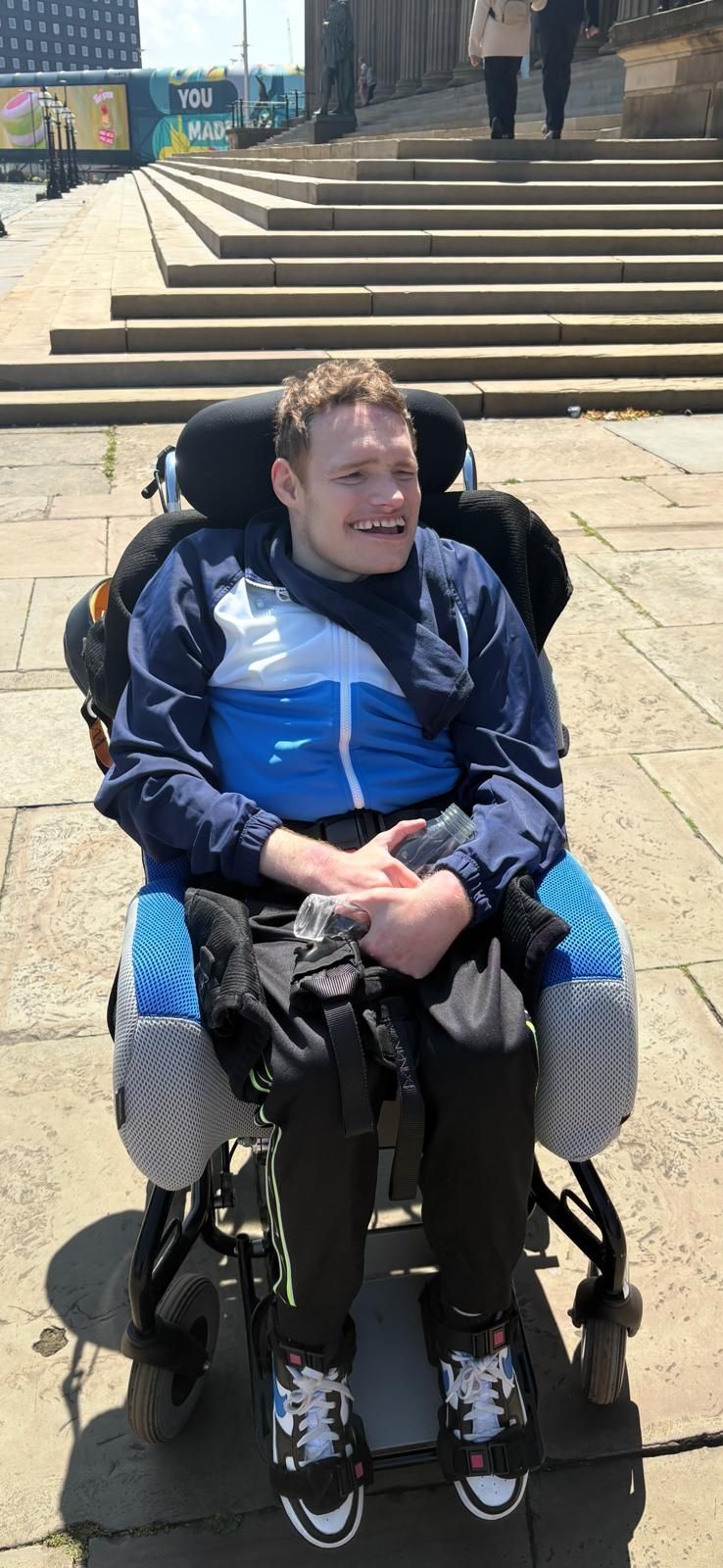 Person in a wheelchair smiling outdoors, wearing blue jacket. Steps and building in background.