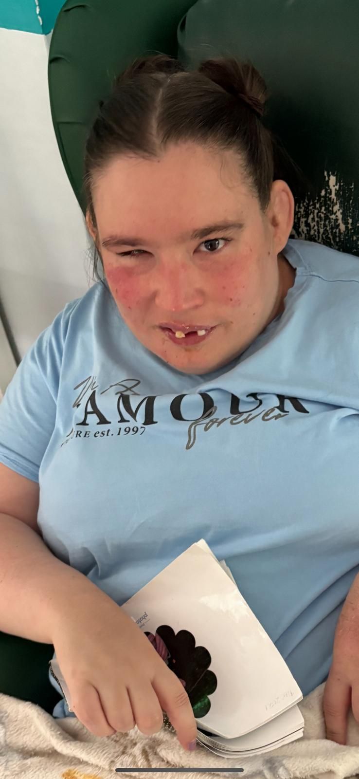 Woman seated in green chair wearing a light blue t-shirt and looking at the camera with a slight smile, holding a craft.