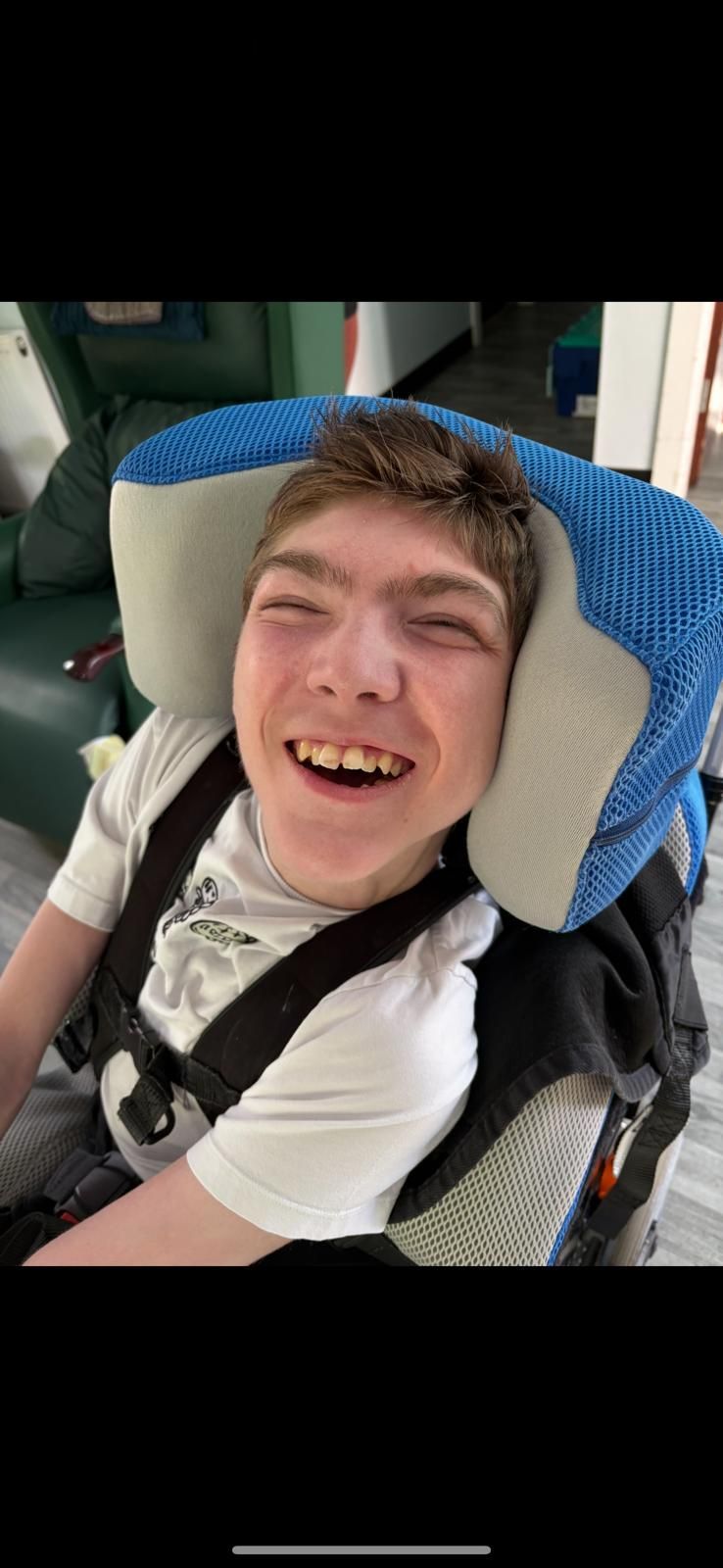 A smiling person in a wheelchair with a supportive headrest indoors, wearing a white shirt, and smiling at the viewer.