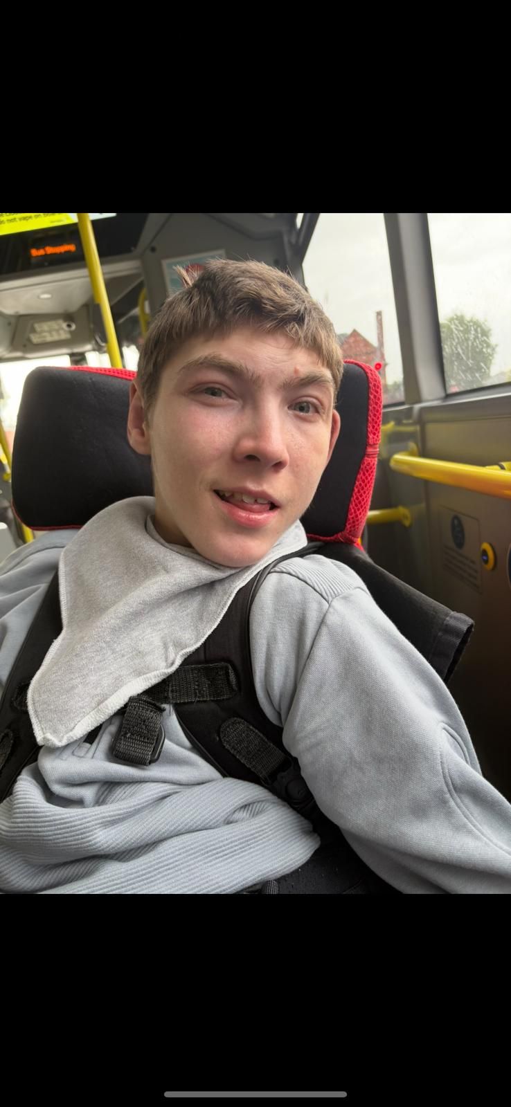A young man with light skin on a bus, smiling. He wears a gray sweater and sits in a seat, with light behind him.