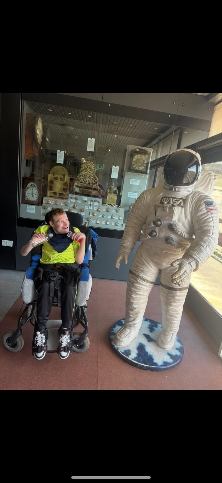 A person in a wheelchair poses with an astronaut statue in front of a display case.