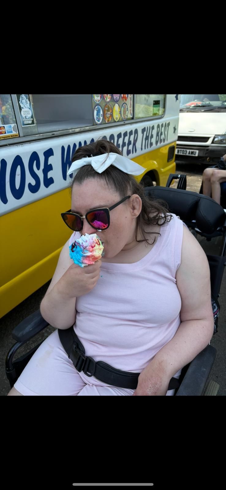 Woman in wheelchair, pink dress, sunglasses, eating colorful ice cream in front of ice cream truck.