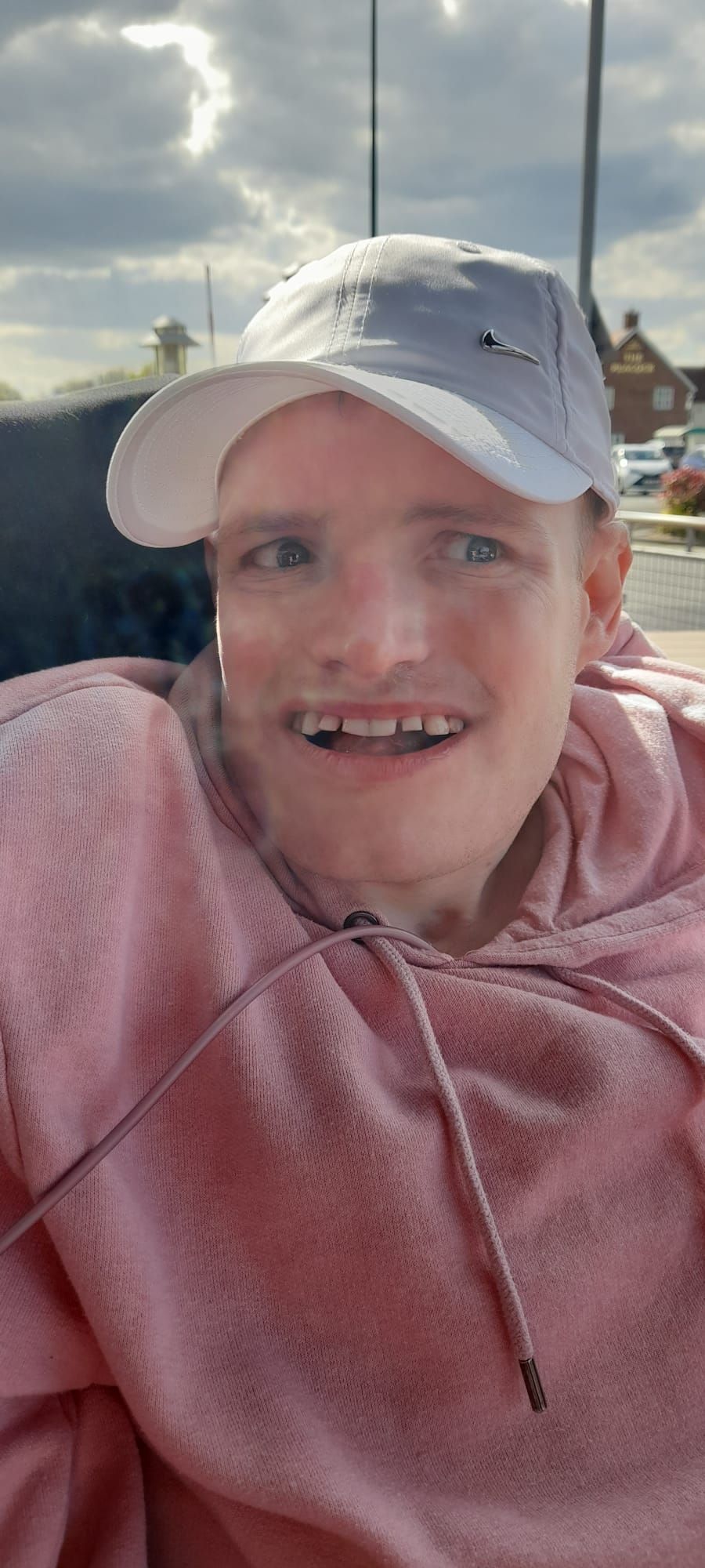 A smiling person wearing a white cap and pink hoodie outdoors. Cloudy sky in background.