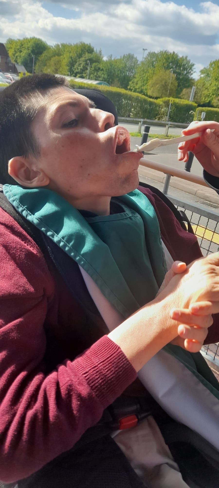Person being fed with a spoon, outdoors. They're wearing a bib and maroon sweater. Lush green trees in background.