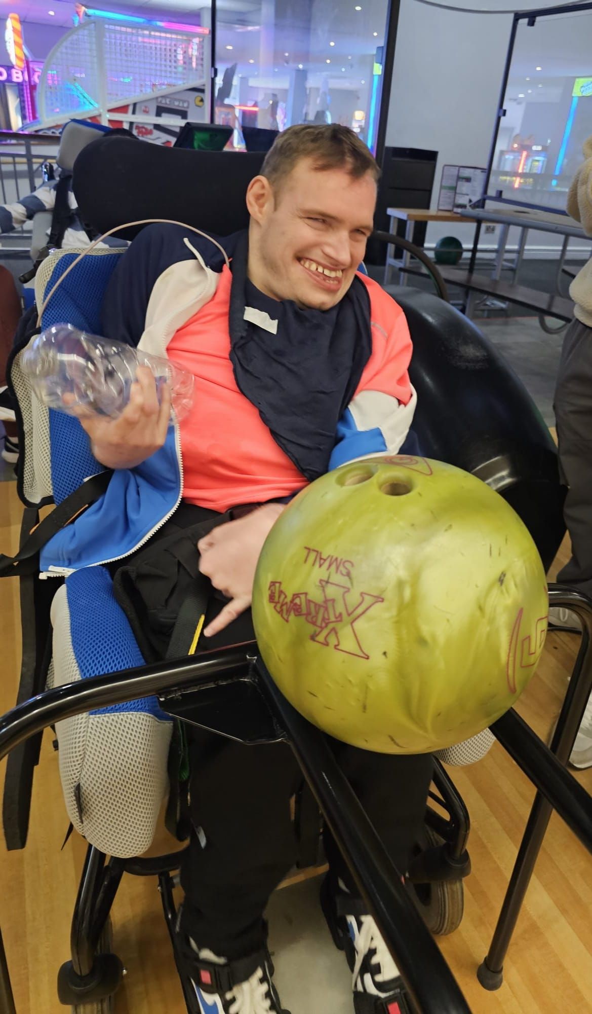 Man in a wheelchair smiles, holding bowling ball and trophy at bowling alley.