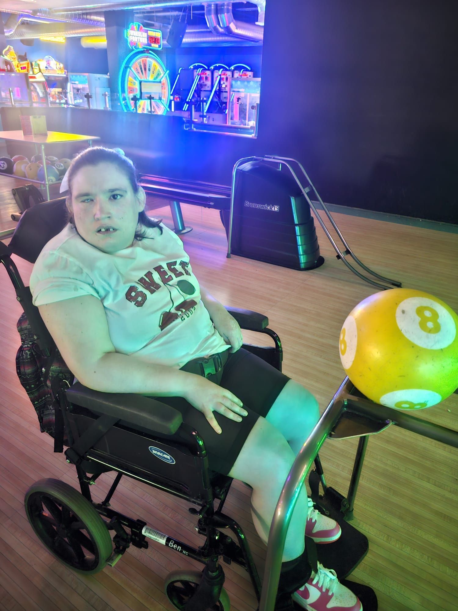 Person in wheelchair at a bowling alley, holding an 8-ball, wearing a t-shirt, and looking forward.