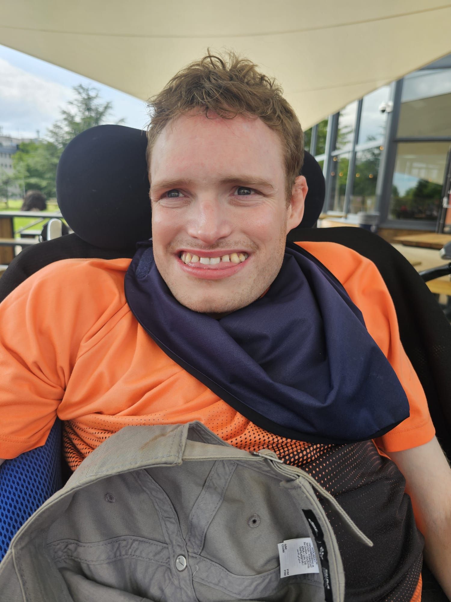 Man in wheelchair smiles, wearing orange shirt, dark blue neck gaiter, and baseball cap outdoors.