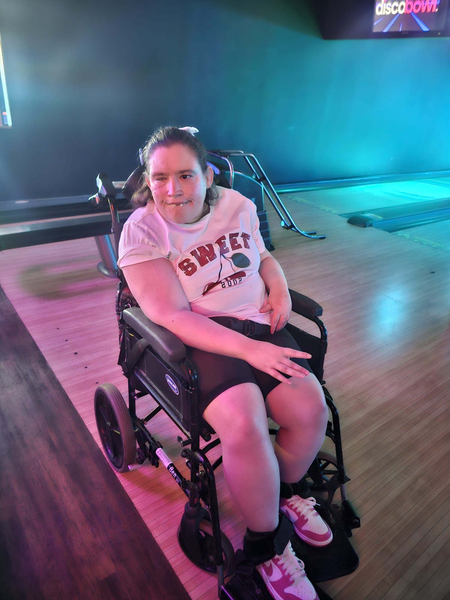 Woman in wheelchair smiles. Wearing white shirt and shorts, in a gym or activity center.