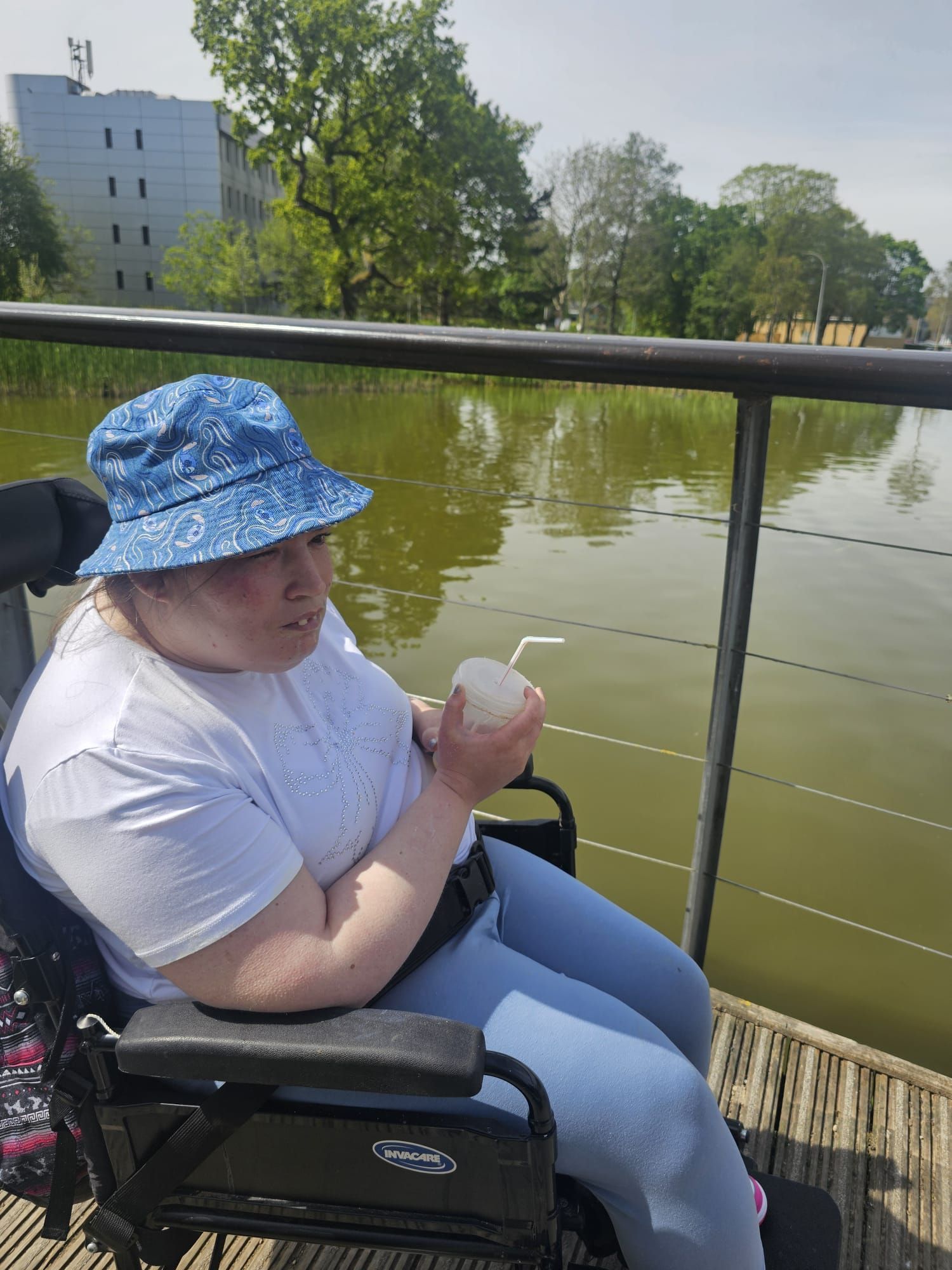 Woman in wheelchair wearing a blue hat, holding a drink near a lake.