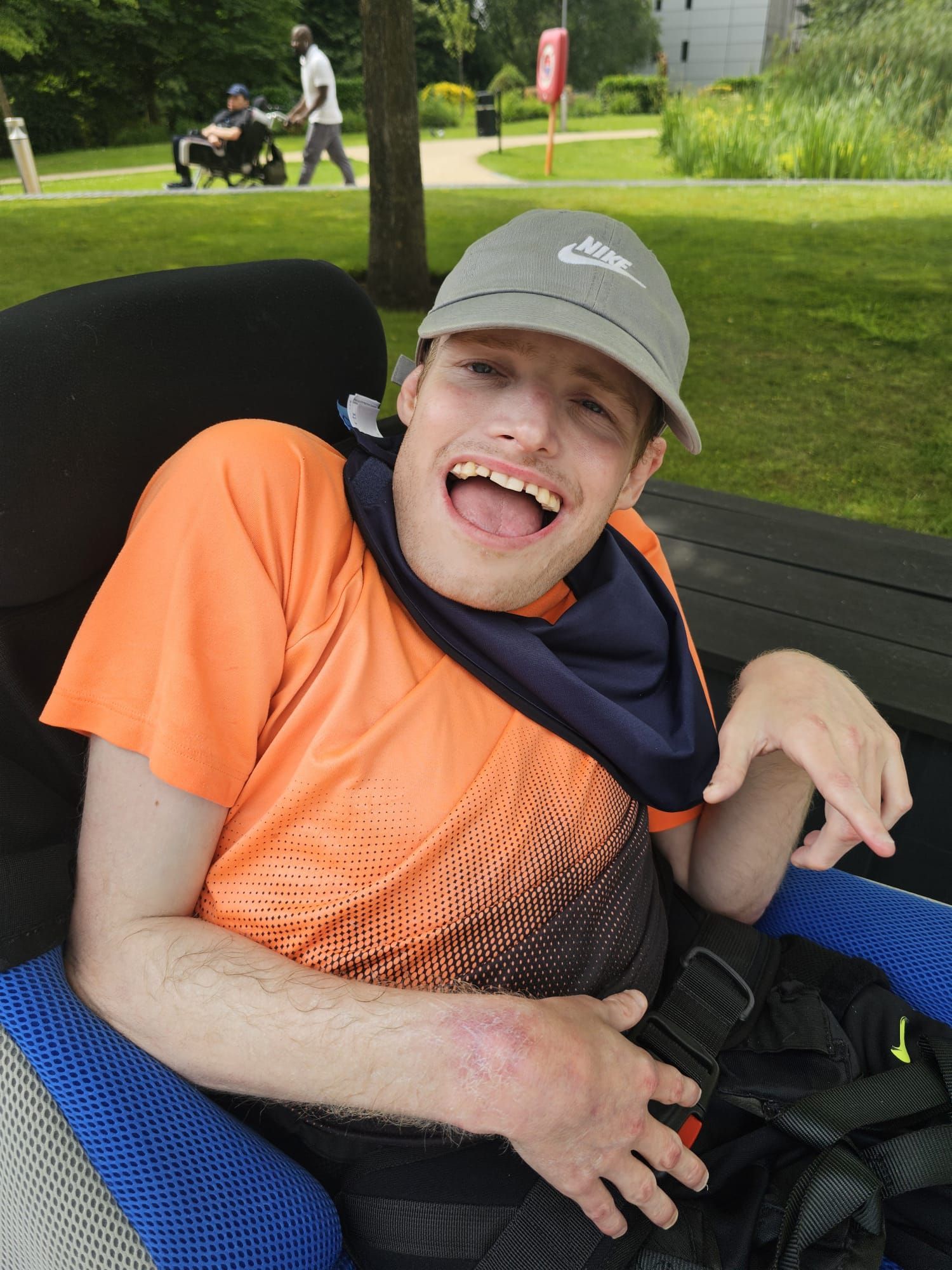 Man in wheelchair laughs, wearing hat and orange shirt, outdoors.
