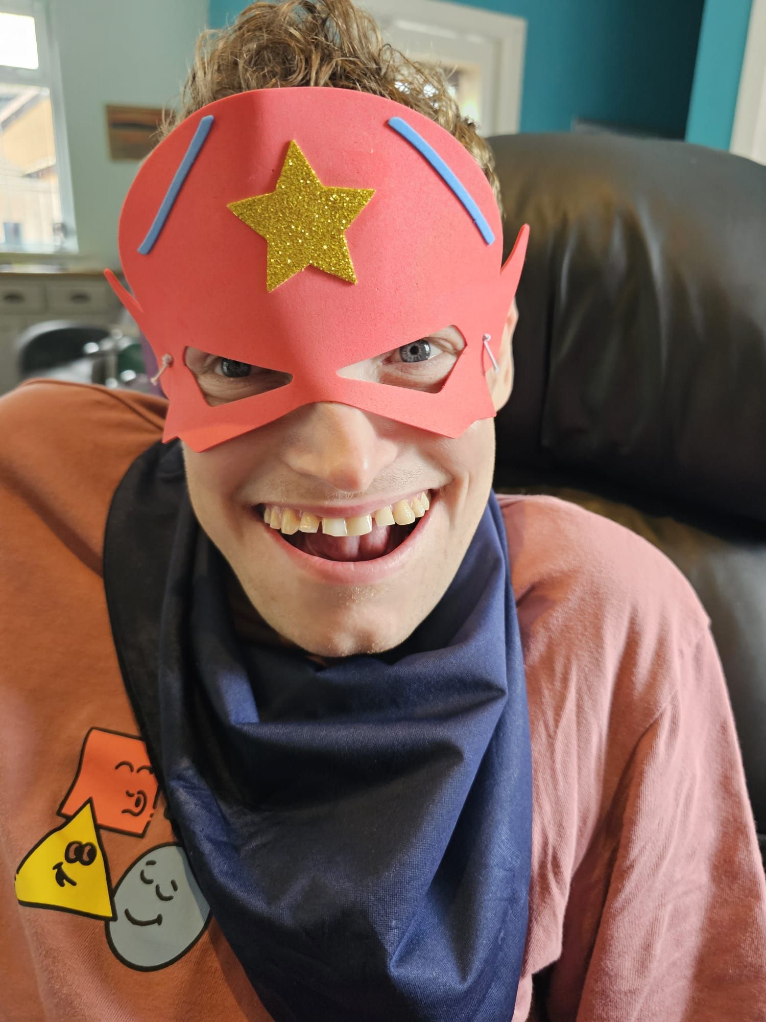 Smiling person in a superhero mask and scarf; wearing a pink shirt. Indoors.