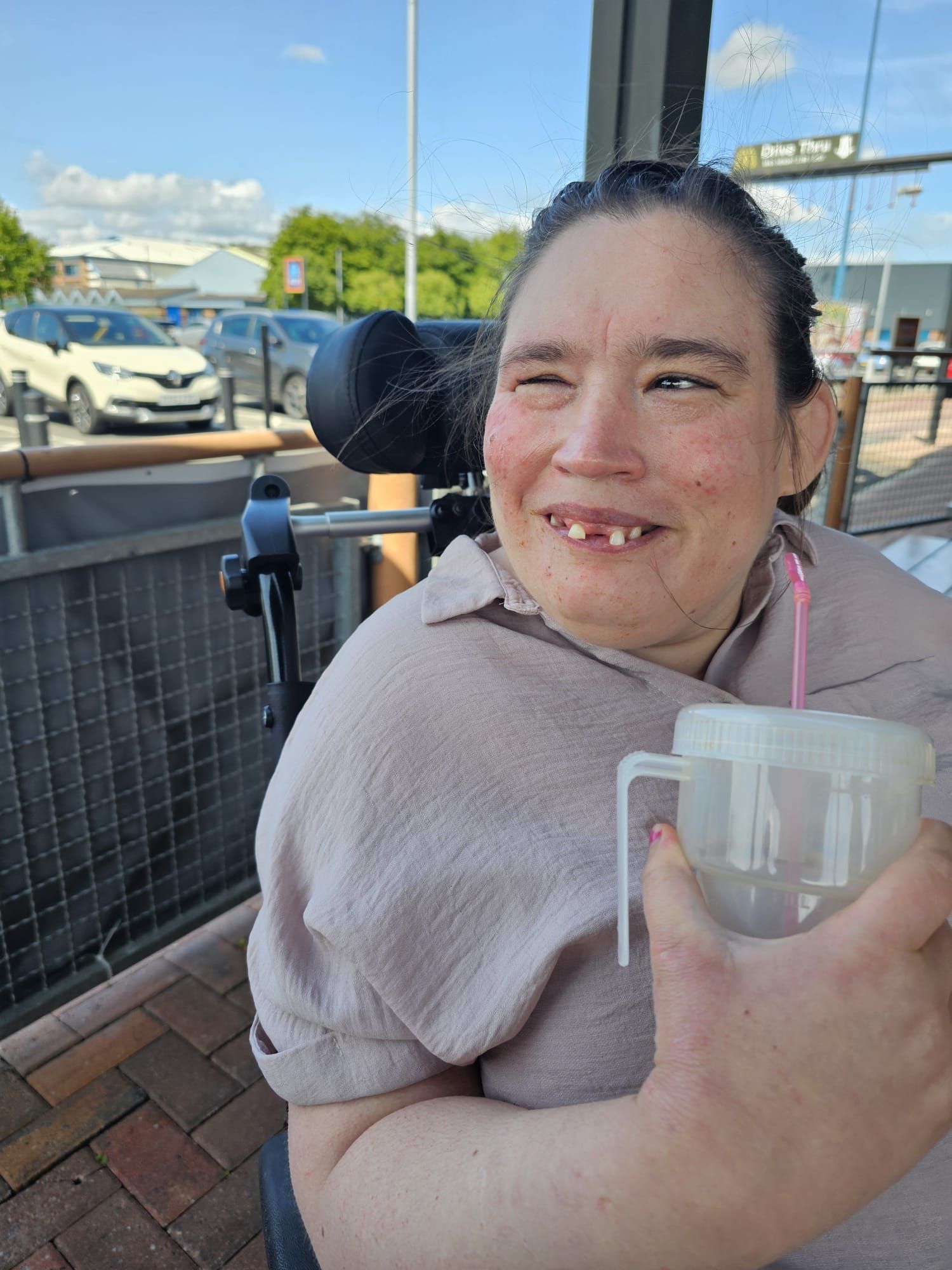 Woman in wheelchair smiling, holding a drink with a straw outside, cars and buildings in background.