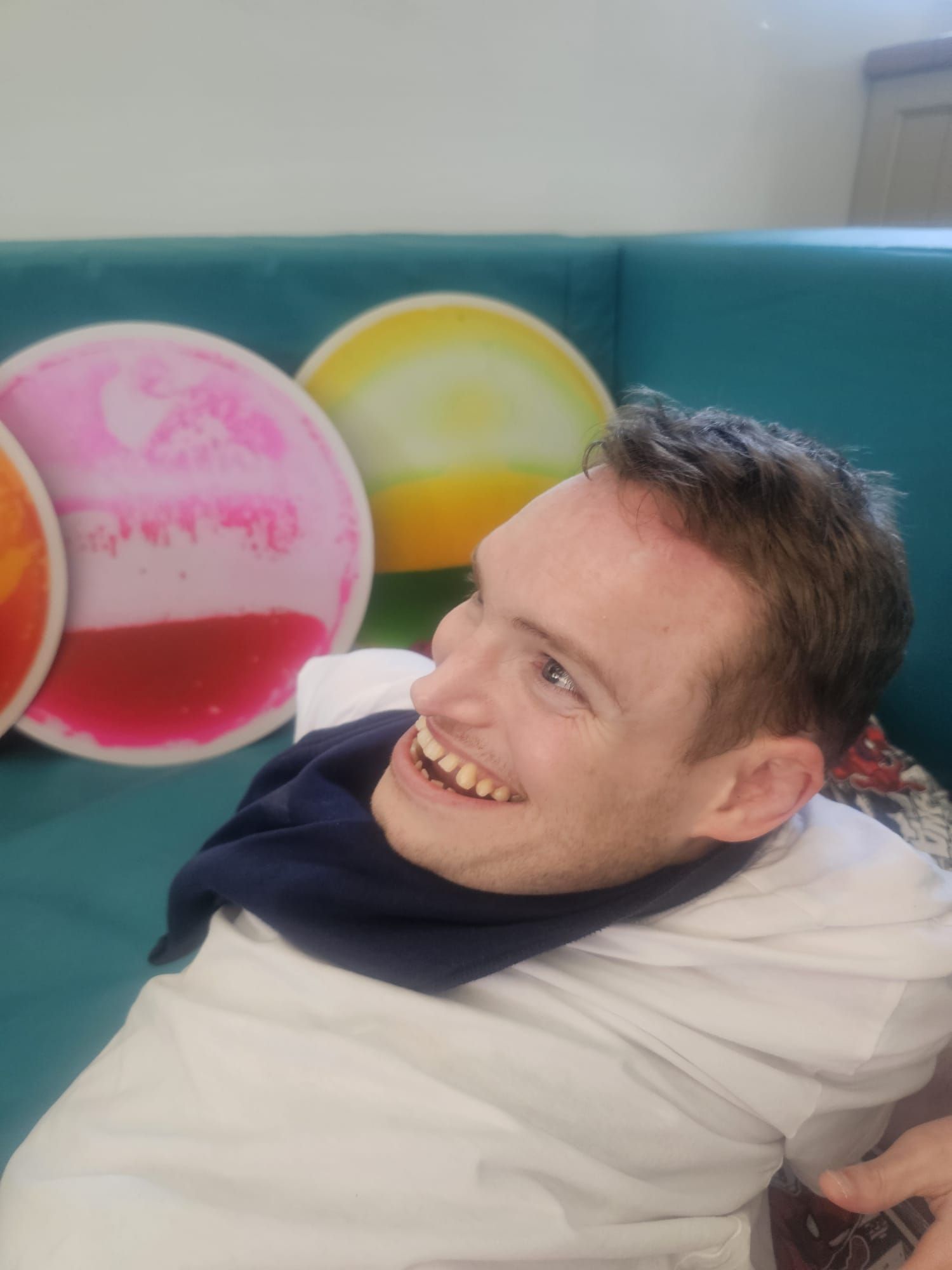 Man smiling, sitting indoors, wearing white shirt and blue neck covering. Colorful circular art in background.
