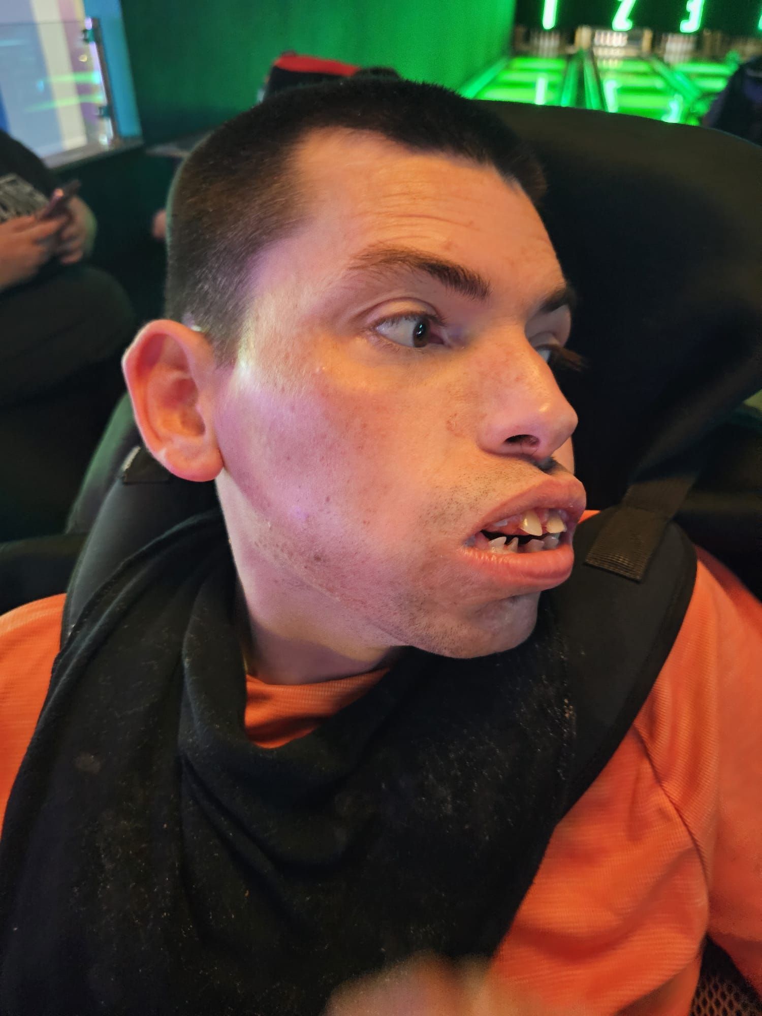 Man with short hair, orange shirt, and black neck support looking to the side in a brightly lit area.