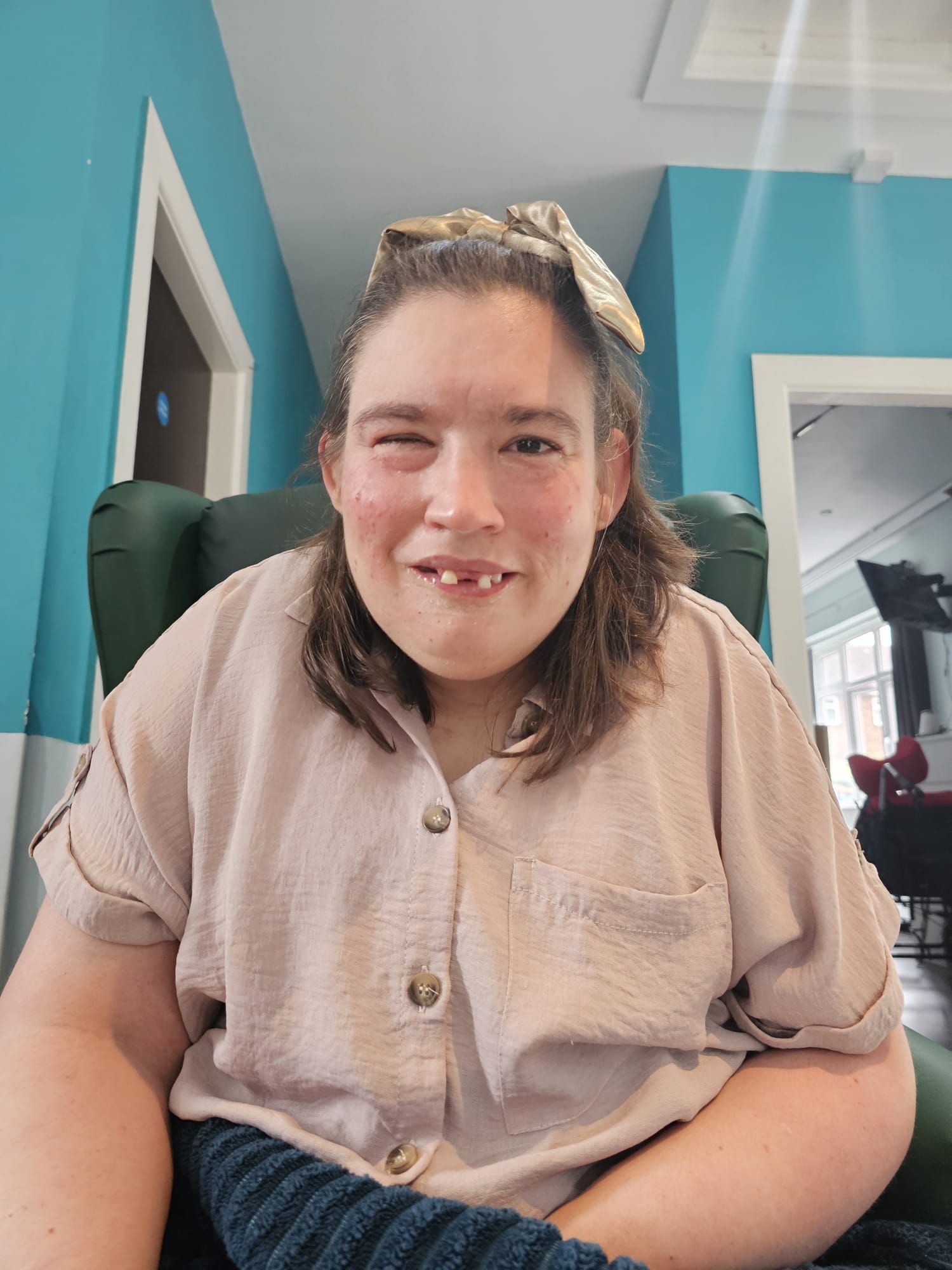 Woman in light pink shirt smiles, seated in a green chair. She has dark hair, and is against a light blue wall.