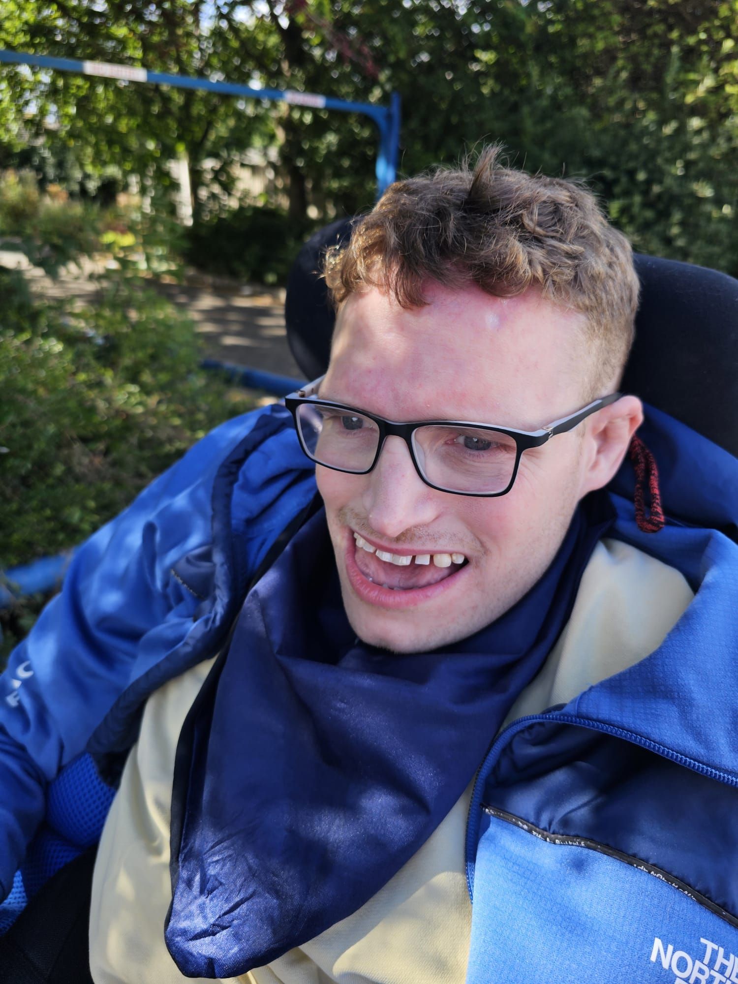 Young man with glasses smiles, seated in a wheelchair outdoors, wearing a blue jacket and scarf.