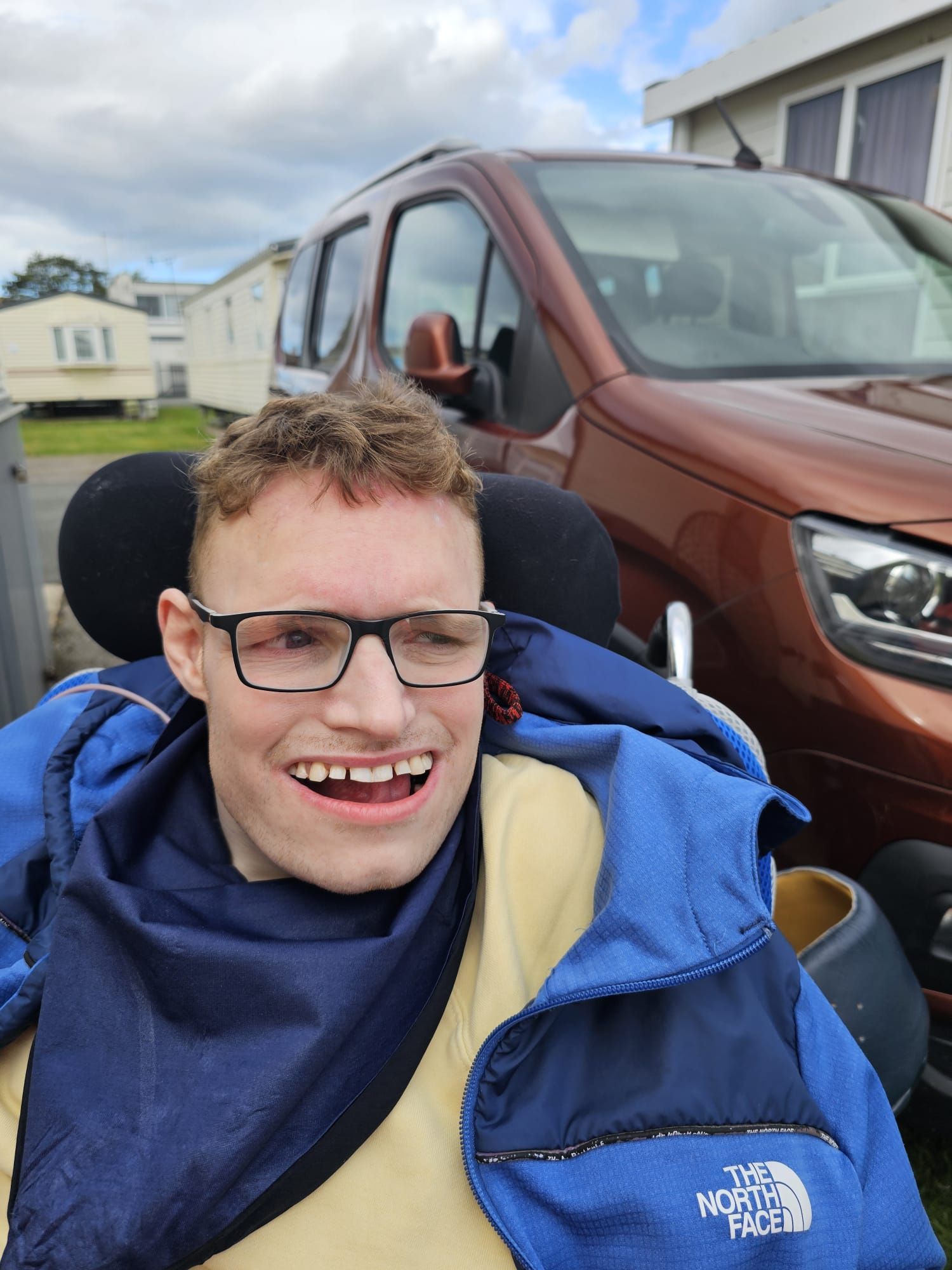 A young man in a wheelchair smiles broadly, wearing glasses and a blue jacket, with a brown van in the background.