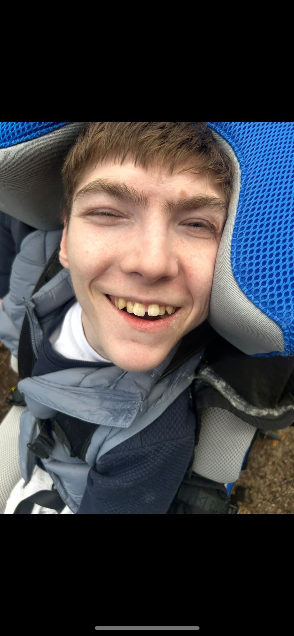 A young man with a warm smile, wearing a jacket with a hood, outdoors.