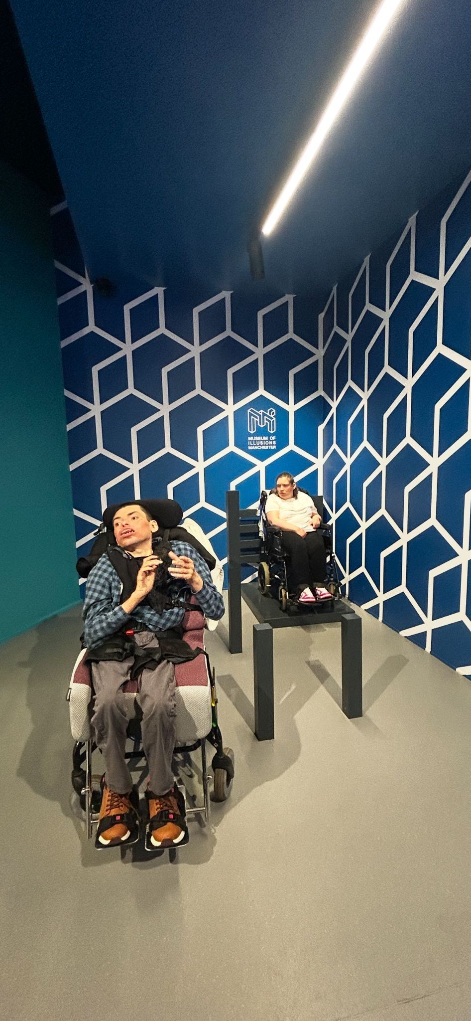 A man in a wheelchair in a room with a geometric pattern on the walls, looking up at a camera.