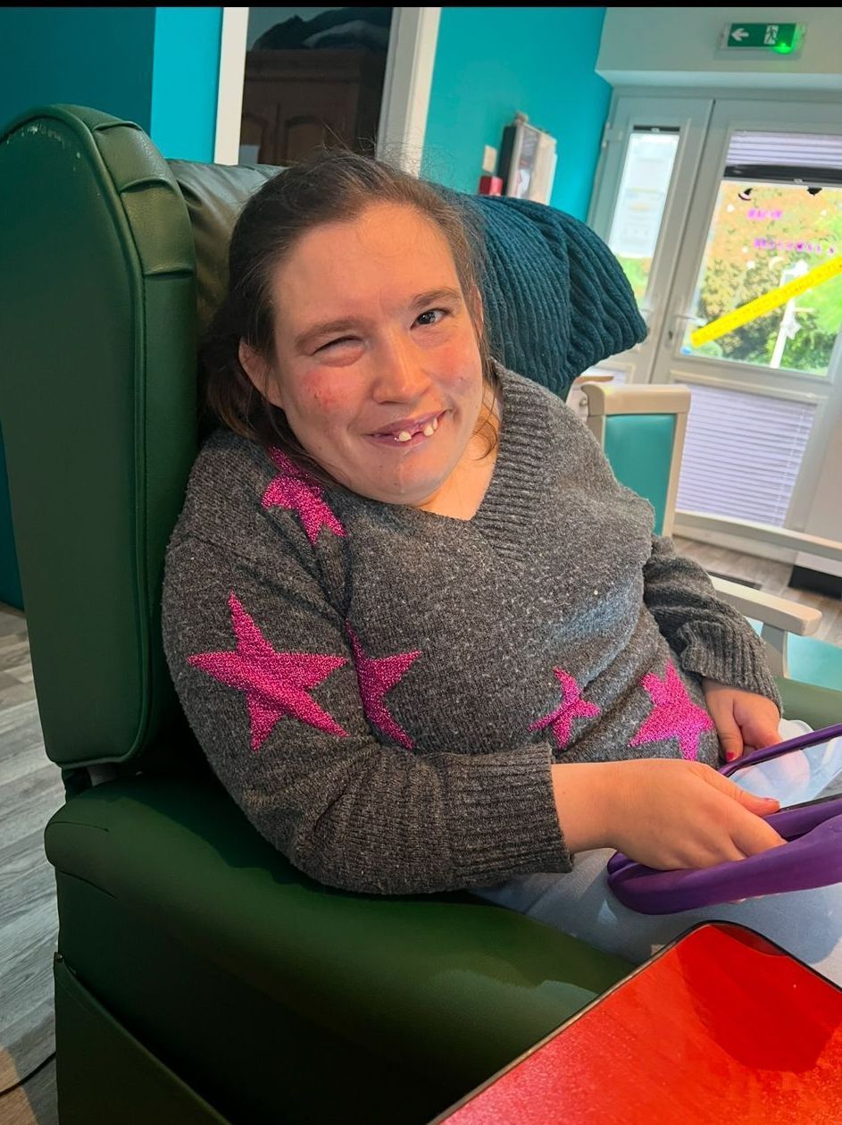 Woman in gray sweater with pink star design smiles, seated in a green chair, inside.