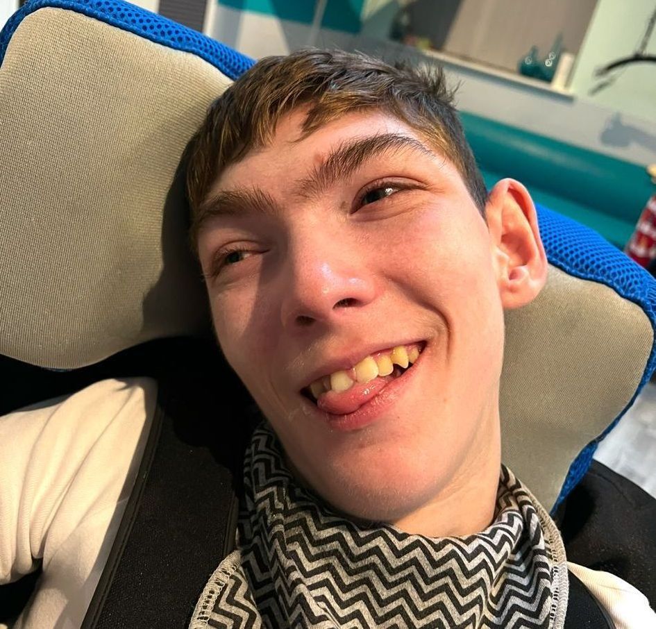 Young man with a warm smile, sticking out his tongue, wearing a scarf, and seated in a chair.