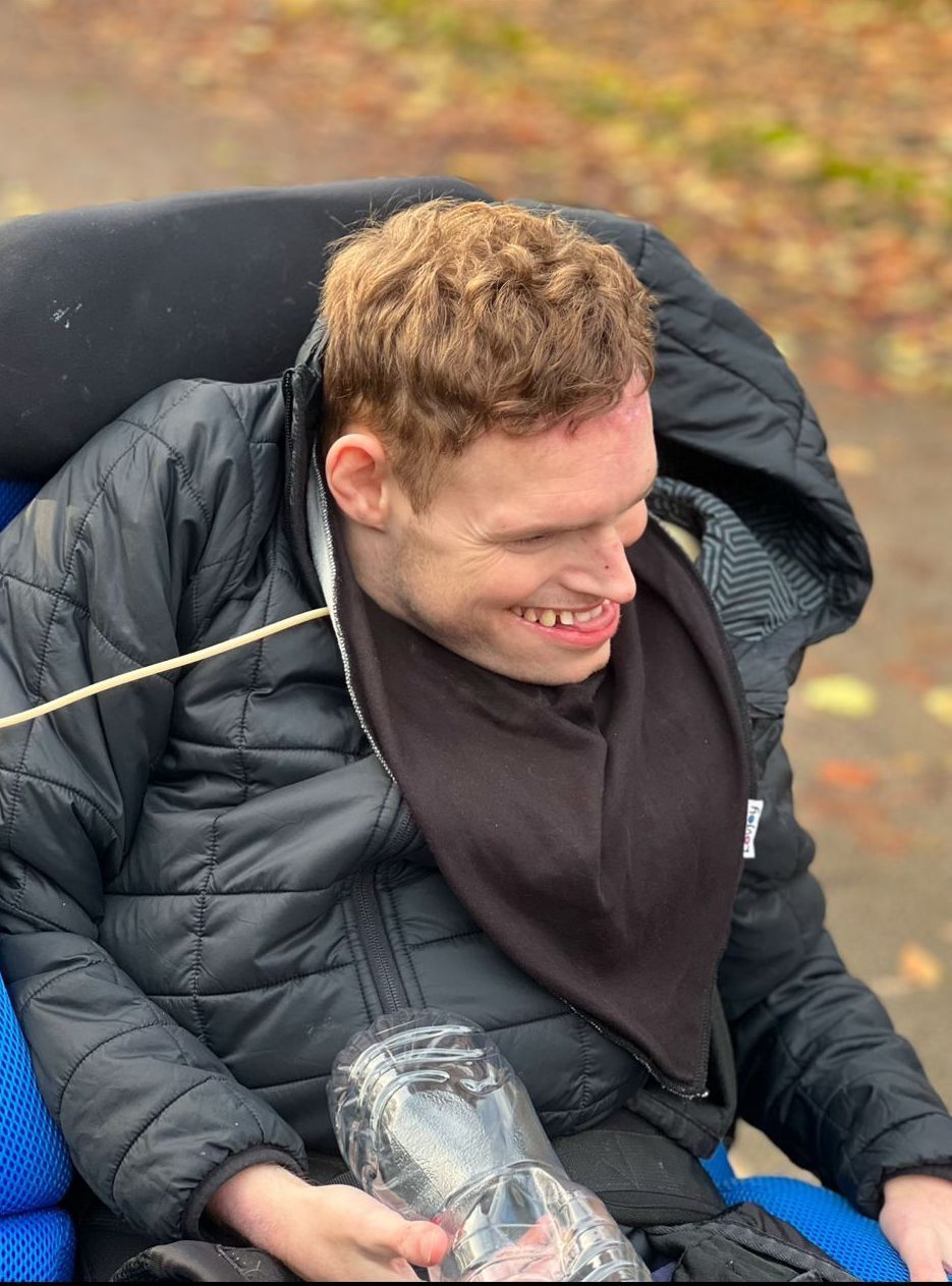 Young man with reddish-brown hair in a wheelchair, smiling. Wearing a black jacket and bib. Outdoors.