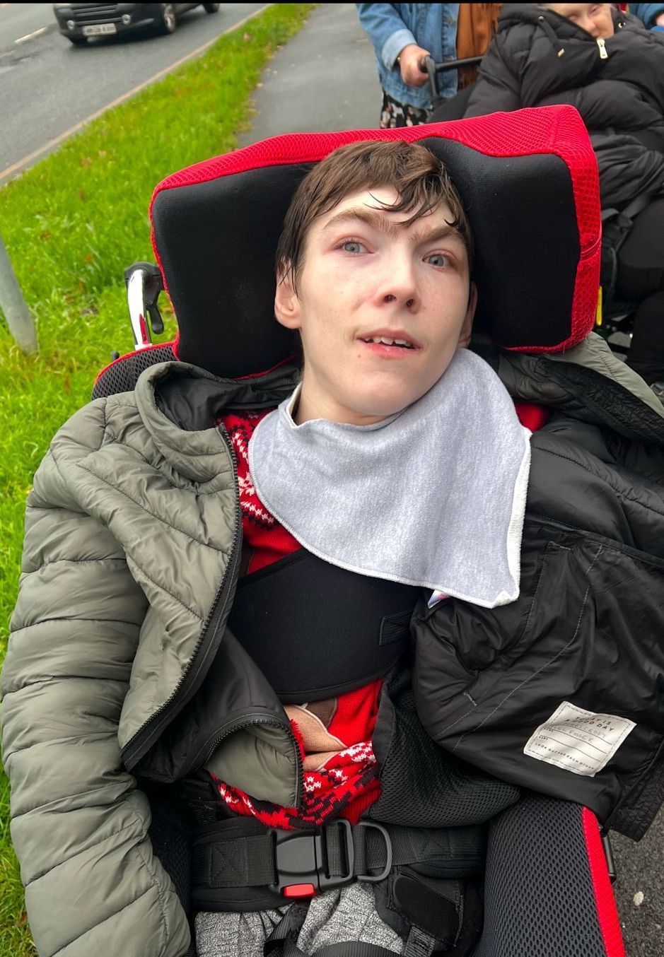 Young person in wheelchair, gray bib, looking at camera, outdoors near green grass.