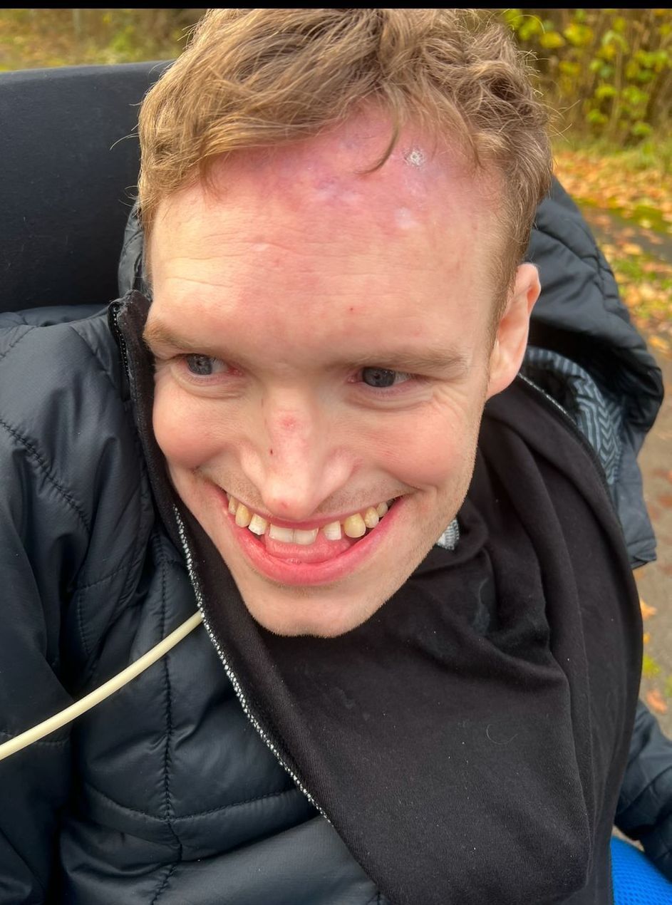 Smiling young man with light skin in a wheelchair; outdoors, black jacket, fall foliage in background.