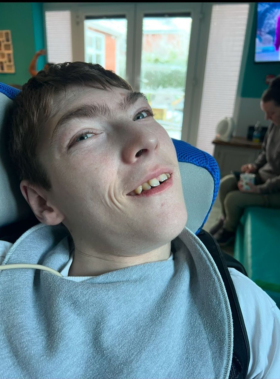 A young man with a slight smile, seated in a wheelchair. He wears a bib and looks towards the viewer.