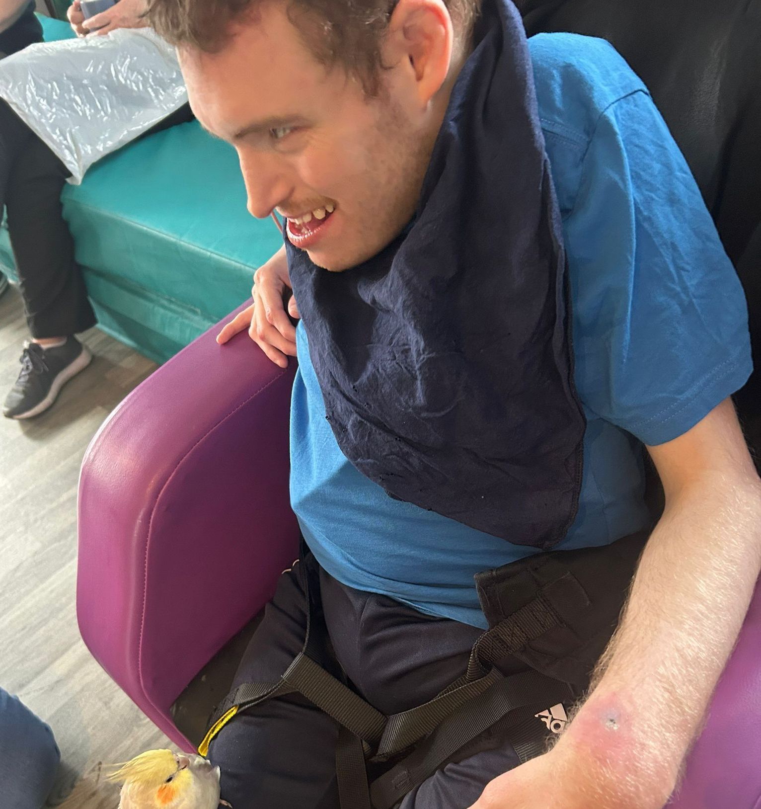 Man in blue shirt smiles, seated in purple chair with black safety straps. Dark blue cloth around neck.