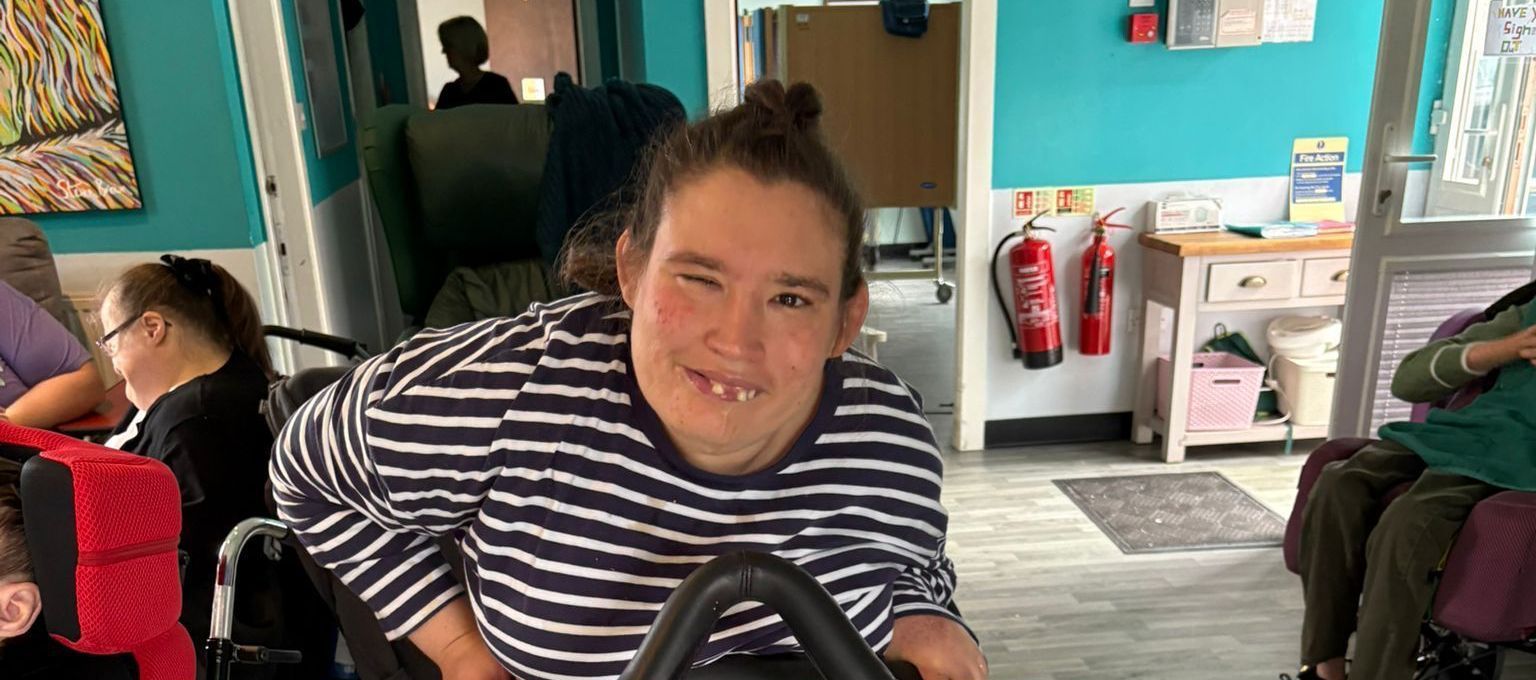 Person in striped shirt smiling in a room, at a care facility. Others are present.