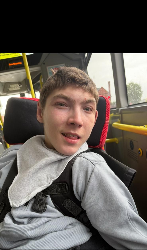 A young person with light skin and brown hair sits on a bus, wearing a gray sweatshirt and a seatbelt.