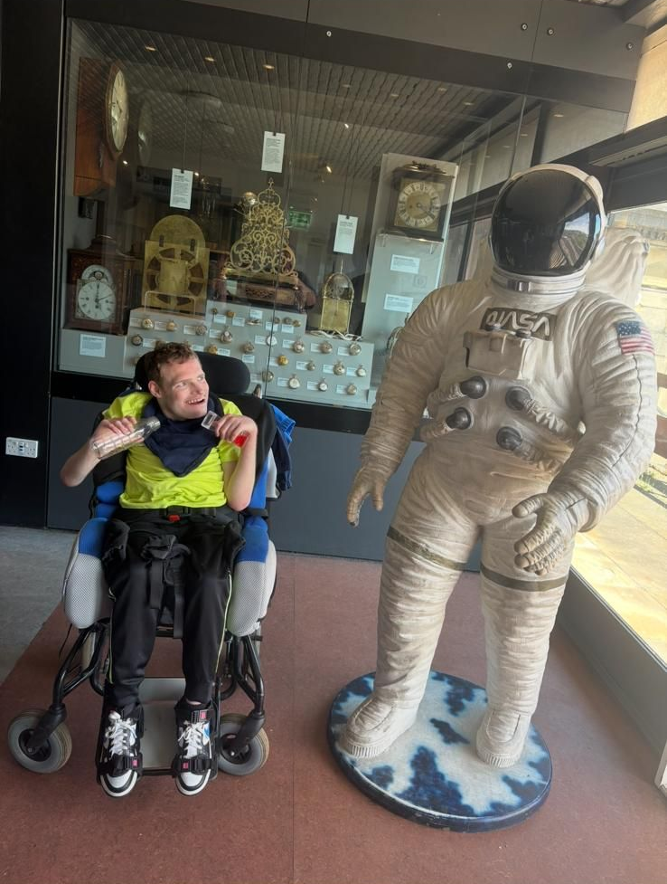 Man in wheelchair next to astronaut statue in museum, clocks in background. Man smiles.
