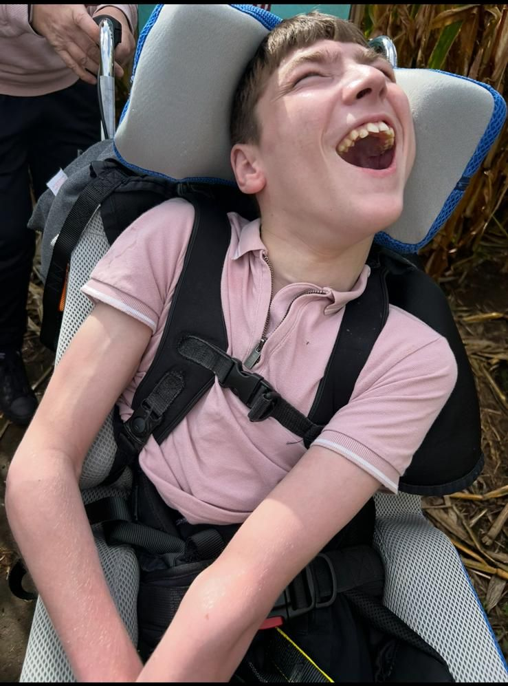 Young person in a wheelchair laughing outdoors, wearing a pink shirt and black straps.
