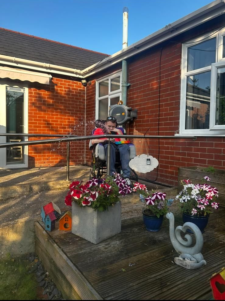 Person in a wheelchair plays a musical instrument on a patio. A brick building and flowers are also present.