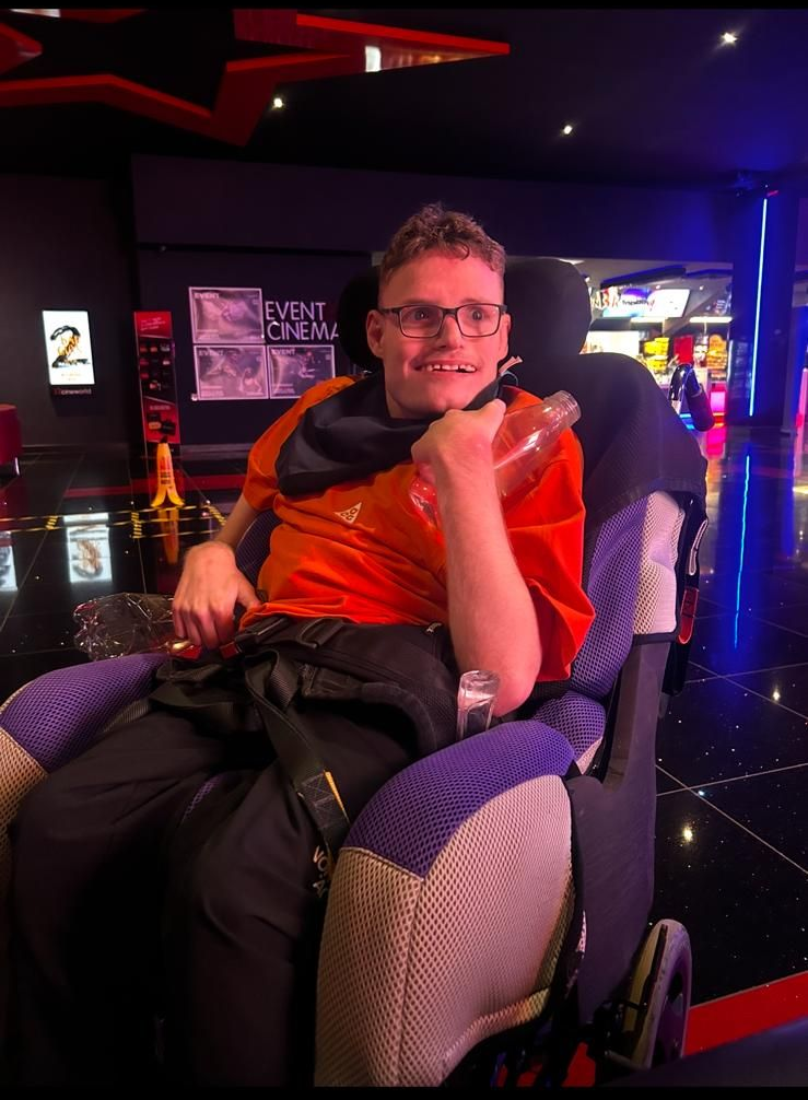 A man in a wheelchair smiles, holding a bottle. He wears glasses and an orange shirt in an arcade.