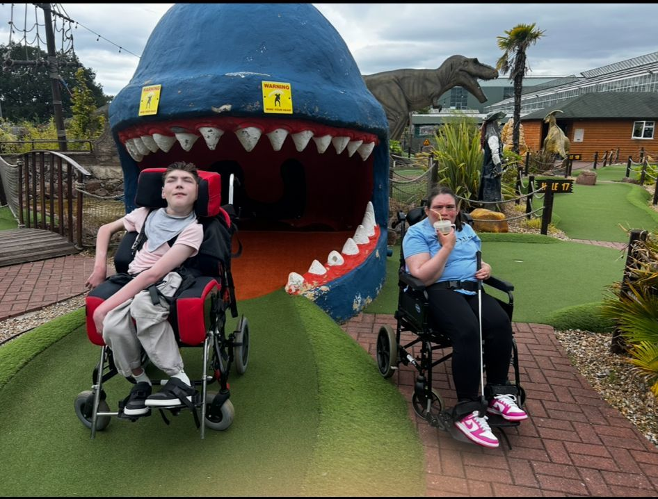 Two people in wheelchairs at a dinosaur-themed mini-golf course, smiling in front of a large, blue dinosaur head.