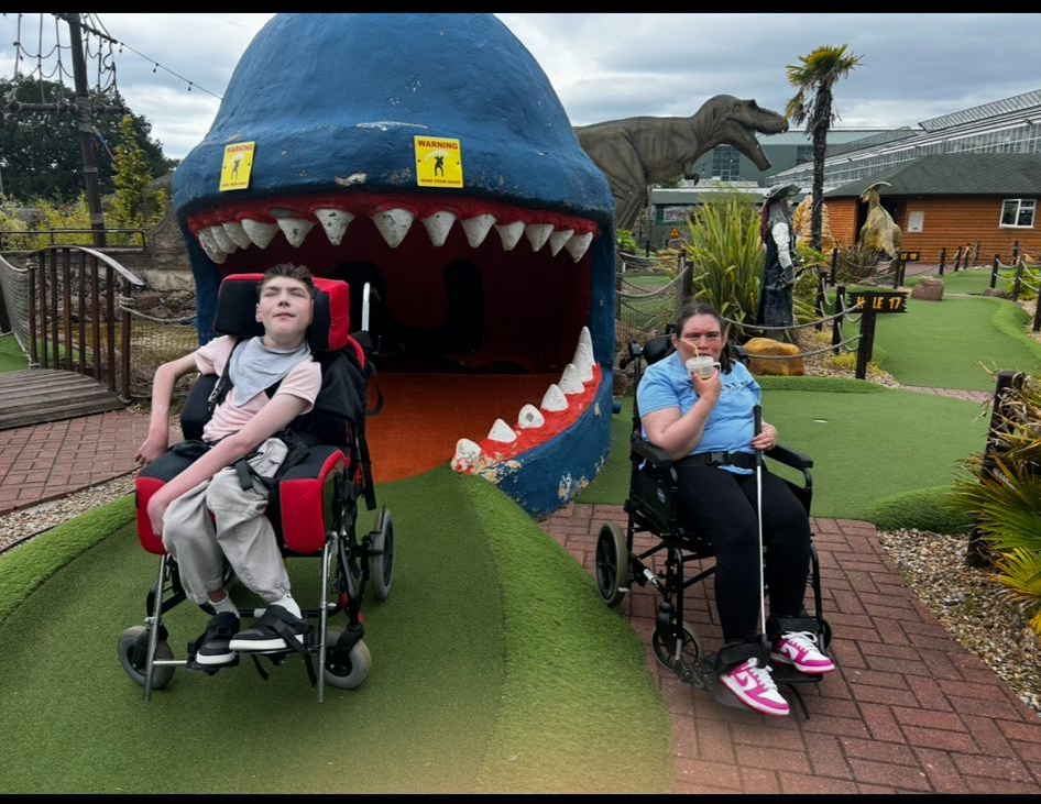 Two people in wheelchairs at a dinosaur-themed mini golf course. One poses, other eats.