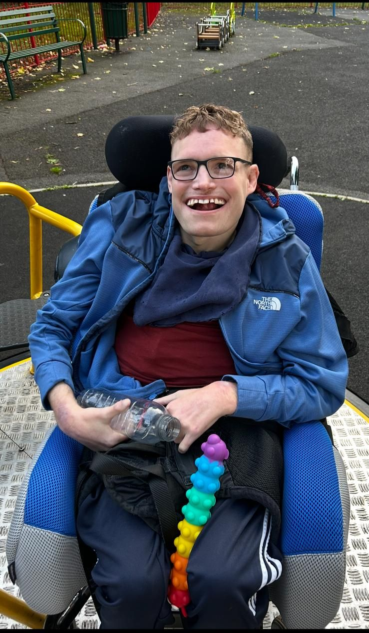 A smiling person in a wheelchair outside, holding a rainbow toy. They wear glasses and a blue jacket.