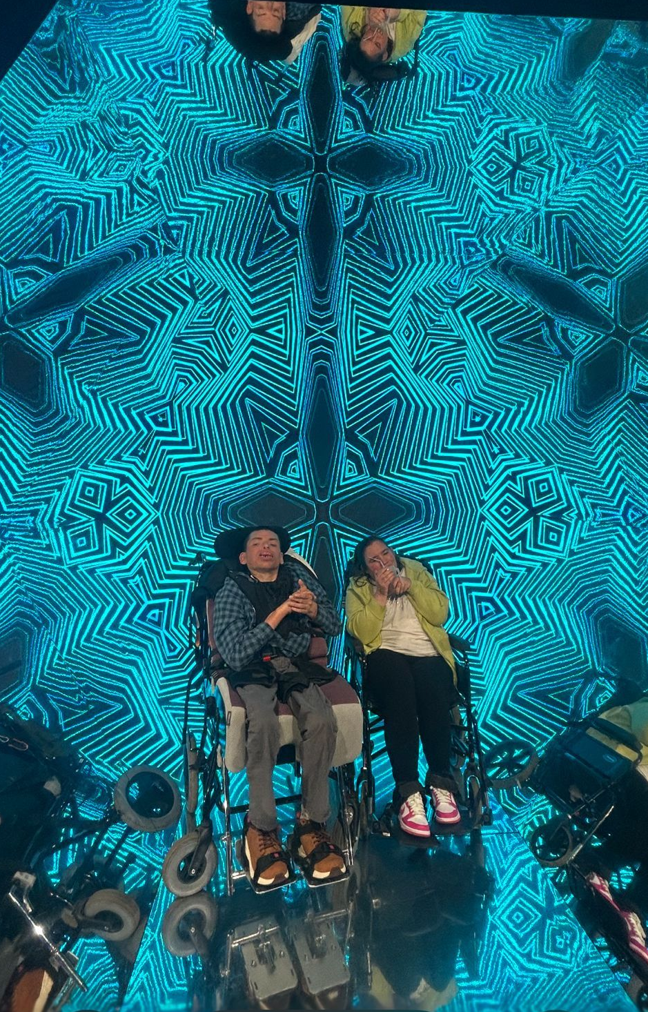 Two people in wheelchairs performing on a stage with a blue kaleidoscope background.