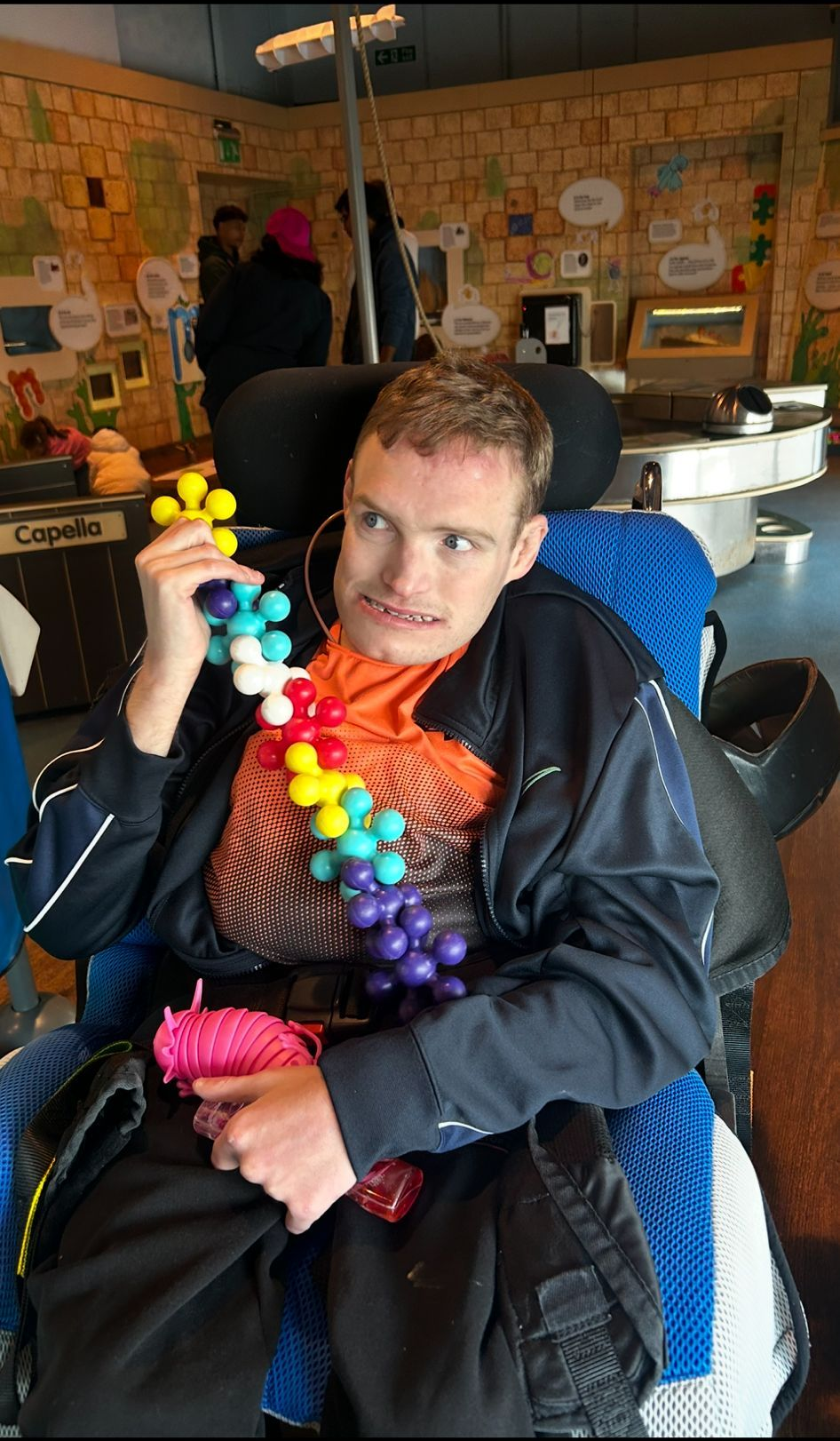 A person in a wheelchair holding colorful beads. Wearing rainbow necklace in an interior space.