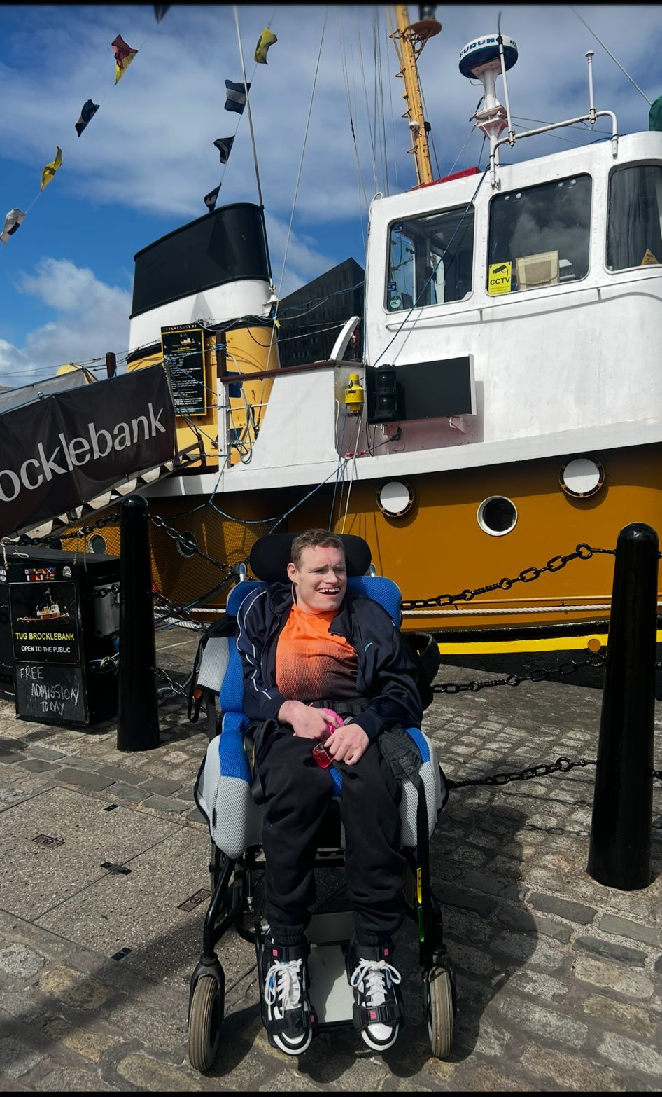 Person in a wheelchair in front of a yellow and white boat. Sunny day, blue sky.