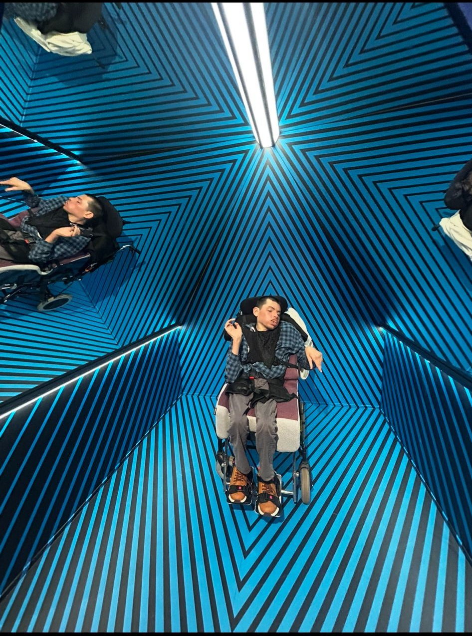 A person in a wheelchair in a blue, patterned hallway with a light above. Two other people are visible on the edges.