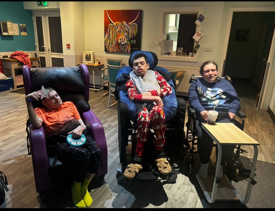 Three people in wheelchairs indoors. One in orange, one in red and white pajamas, and one in blue. Cozy setting.