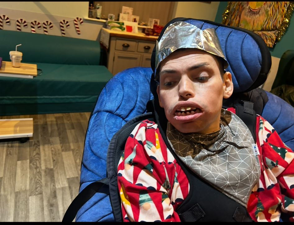 Person in wheelchair wearing a foil crown and holiday shirt, smiling, indoors.