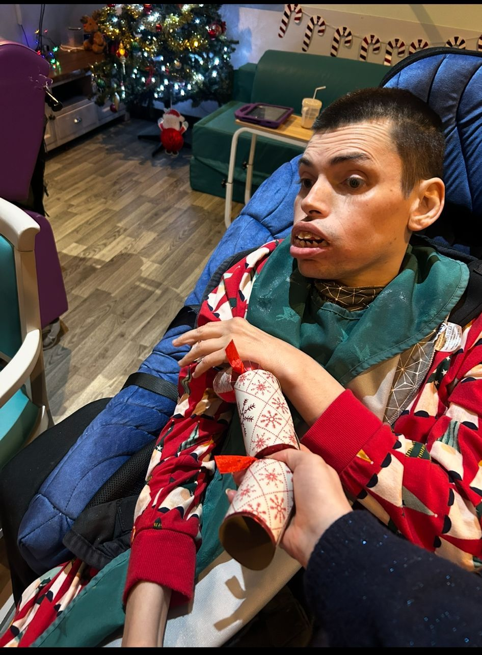 Man in Christmas attire holding a cracker; decorated room with a tree in the background.