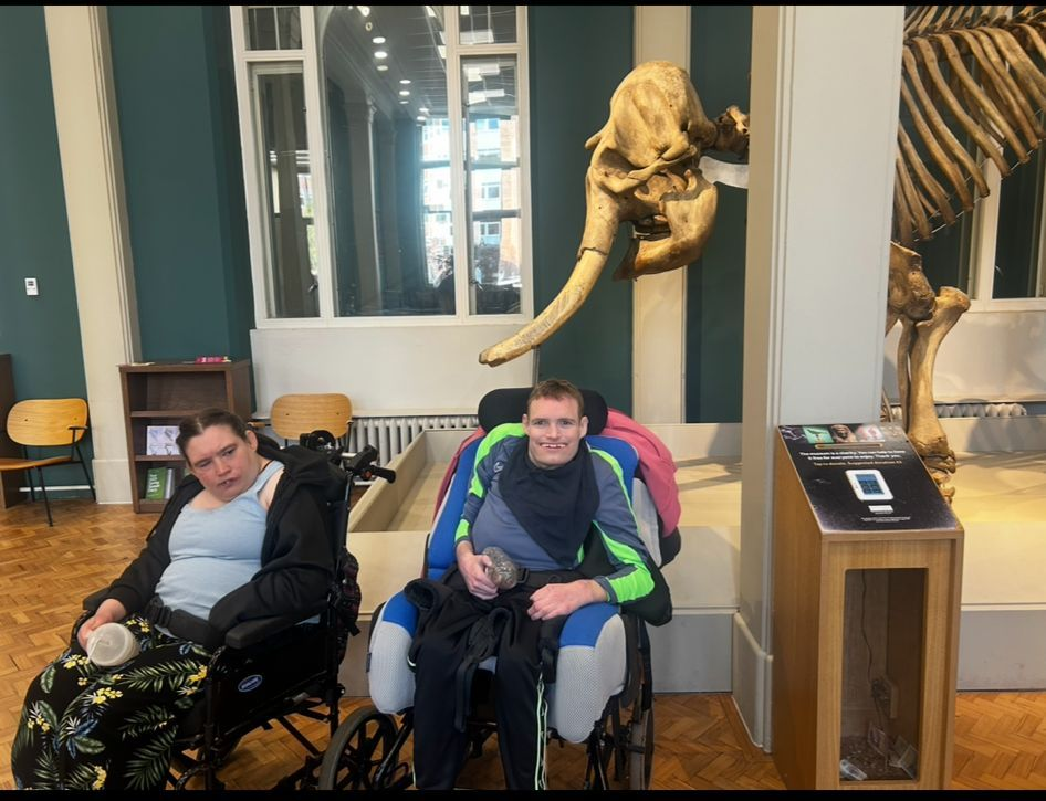 Two people in wheelchairs at a museum, looking at a large bone display.