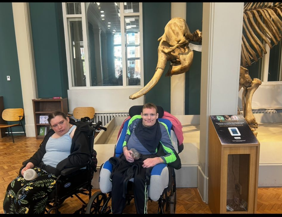 Two wheelchair users pose at a museum exhibit, an elephant skeleton and display case in the background.