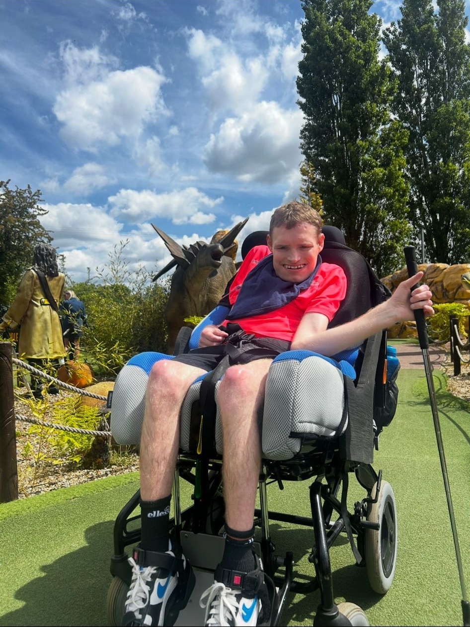 A person in a wheelchair outside on a sunny day; holding a walking stick.
